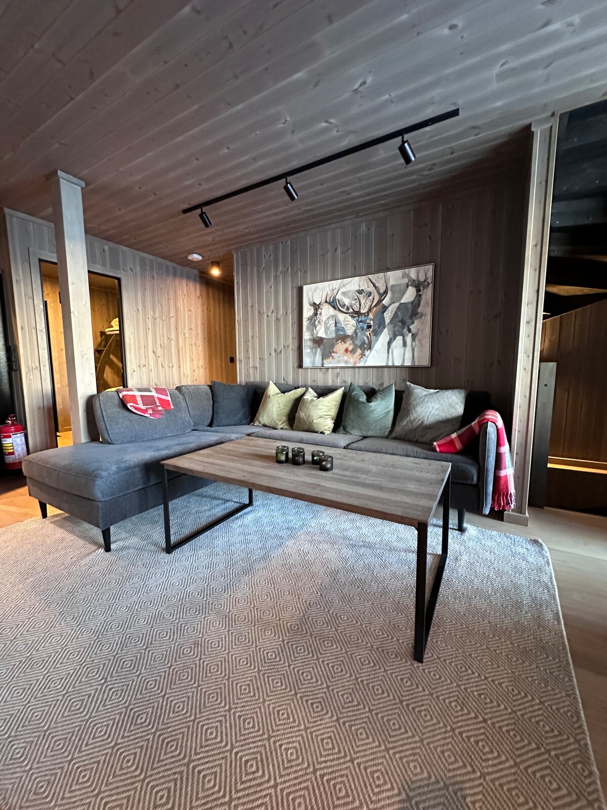 A comfortable seating area is presented with a sectional grey sofa adorned with decorative pillows. A wooden coffee table sits at the center, complemented by a patterned area rug beneath. Wall art featuring wildlife decorates the wall, and a staircase is visible in the background.