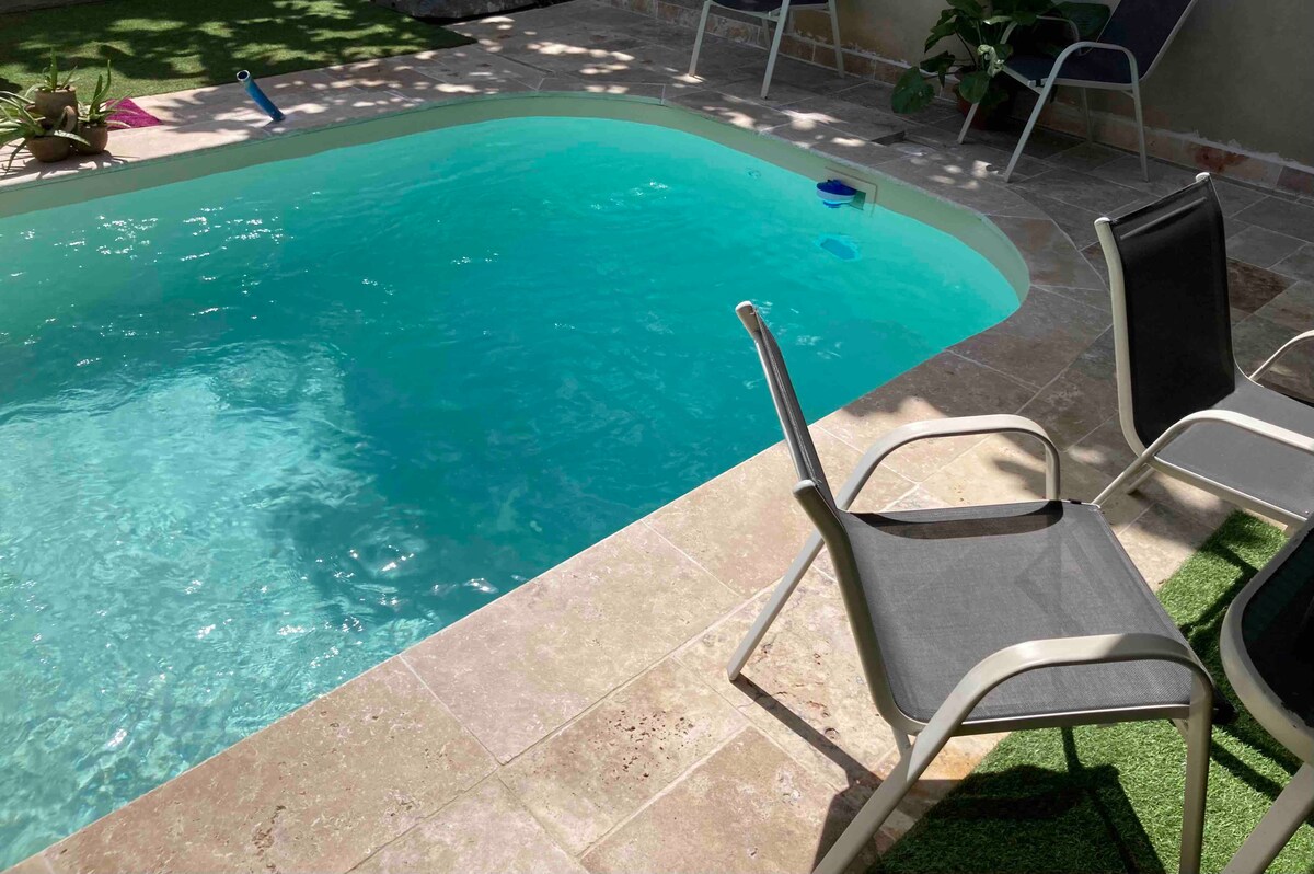 A refreshing pool is surrounded by a stone patio, featuring two modern lounge chairs positioned nearby. Sunlight reflects off the turquoise water, while greenery borders the area, creating a serene outdoor space.