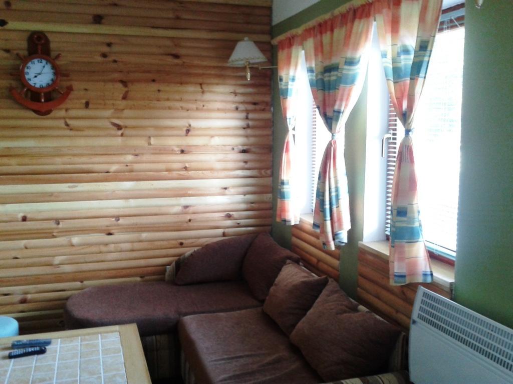 A cozy sitting area features a brown sectional sofa adorned with plush cushions. Light filters through two patterned curtains, which frame wood-paneled walls. A round table is positioned in front of the sofa, and a wall clock adds a functional touch to the space.