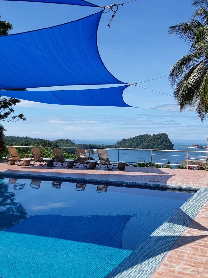 A refreshing swimming pool reflects the clear blue sky, surrounded by lounge chairs positioned for sunbathing. Shade sails extend overhead, providing comfort from the sun, while a stunning view of the ocean and lush landscape is visible in the background.