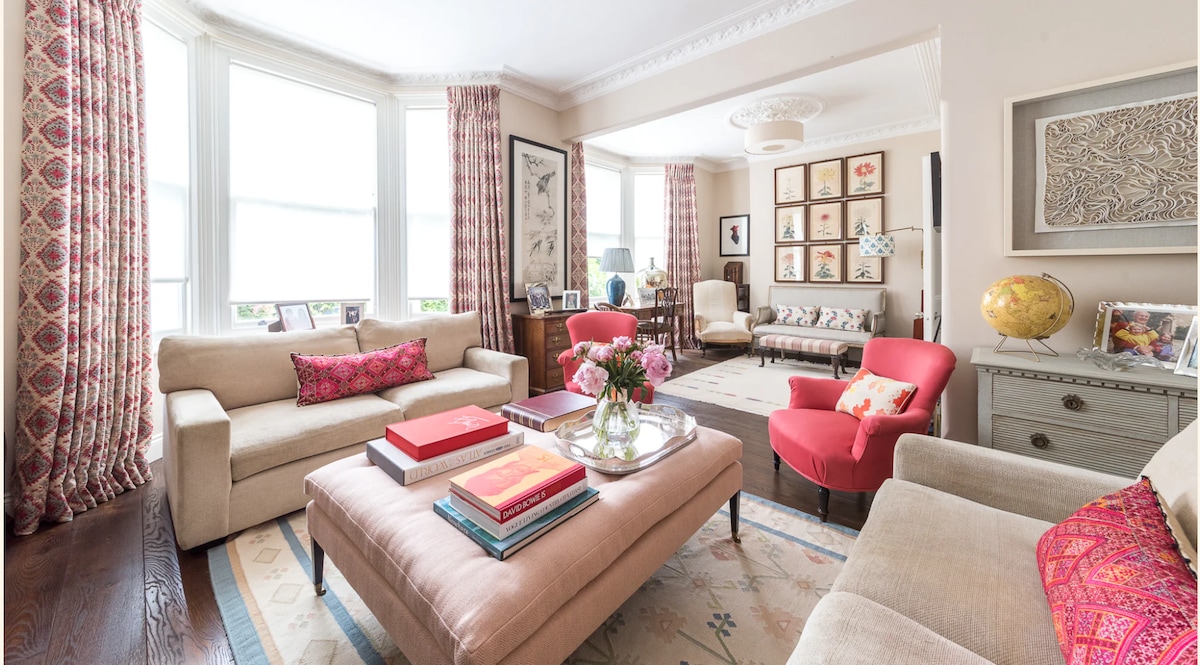 A bright living room features large windows dressed with patterned curtains, allowing natural light to fill the space. Two sofas are positioned around a central coffee table, adorned with books and flowers. Colorful art frames line the walls, adding visual interest to the inviting atmosphere.