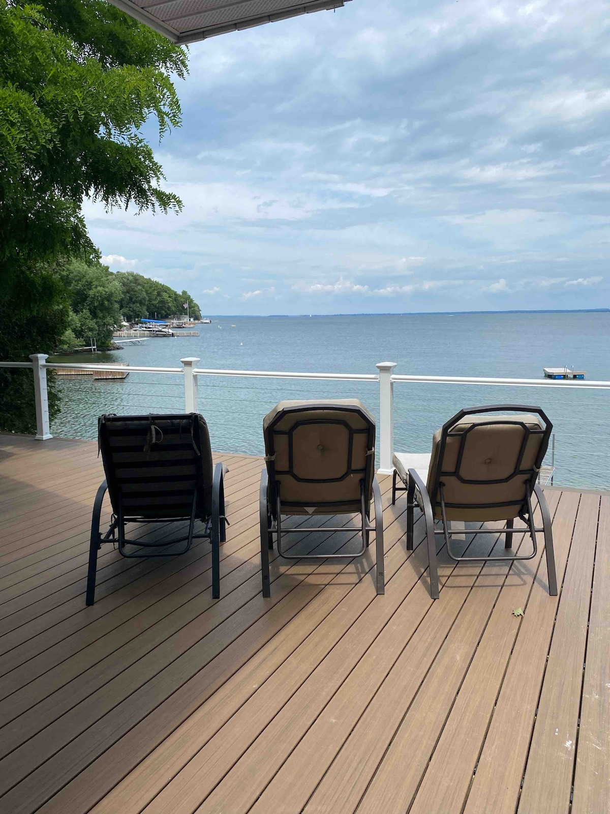 Three lounge chairs are positioned on a wooden deck, facing a calm waterfront scene. The view includes the expansive lake, with a gentle shoreline visible on the left and boats bobbing in the distance under a partly cloudy sky.