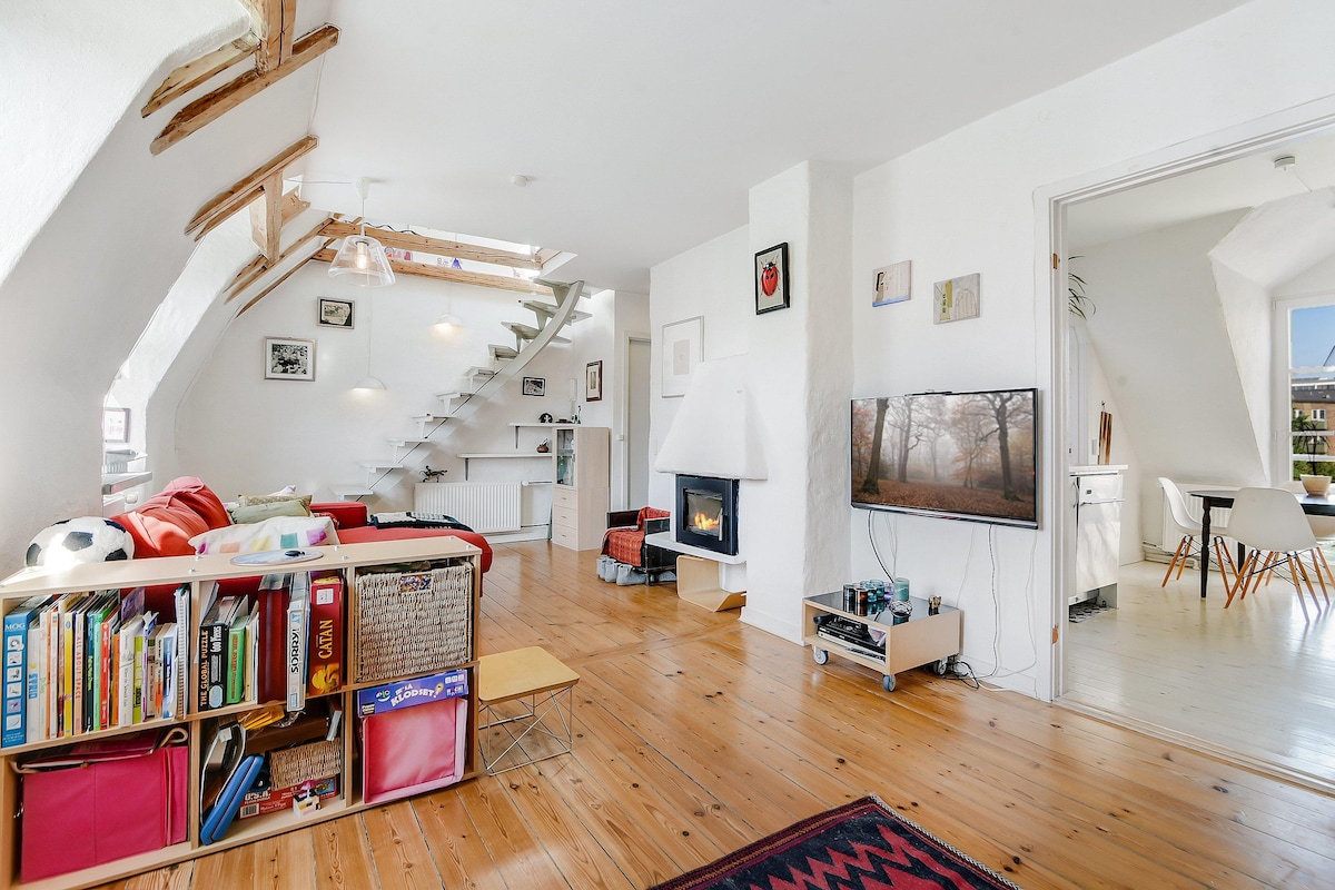 A bright living space is characterized by wooden flooring and open architecture. A cozy fireplace is positioned next to a modern television. A colorful couch faces the fireplace, surrounded by shelves filled with books. Natural light filters through windows, enhancing the atmosphere.