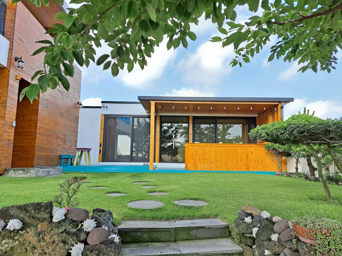 The exterior of the rental house features a welcoming porch area and expansive glass windows, allowing natural light to fill the space. Lush green grass surrounds the home, with stepping stones leading to the entrance, framed by trees and shrubs.