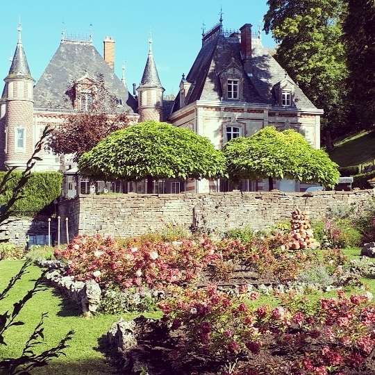 The image captures a historic chateau with distinctive spires and ornate architecture, set against a clear blue sky. Lush greenery and well-maintained flower beds with vibrant blooms create a picturesque garden in the foreground, enhancing the property’s serene atmosphere.