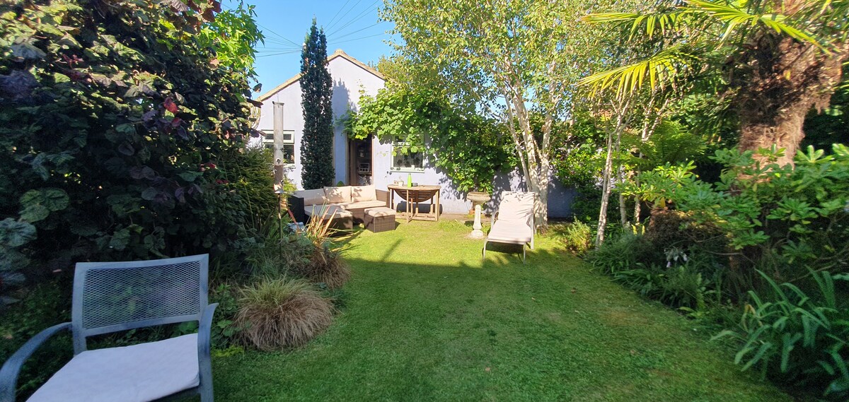 A green garden area is shown, featuring a lawn surrounded by diverse plants and trees. Two lounge chairs are positioned on the grass, and a wooden table with chairs is visible under the shade of trees. A sense of tranquility is conveyed by the lush surroundings.