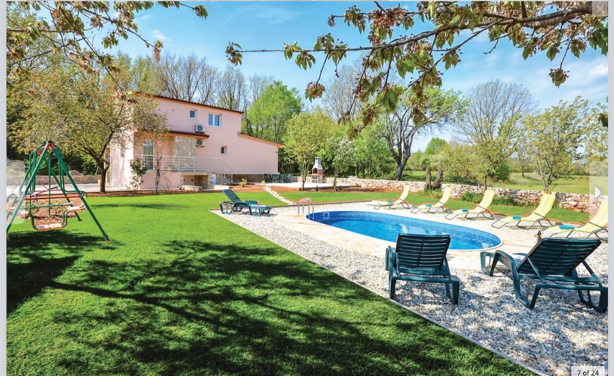 A spacious outdoor area features a blue circular pool surrounded by sun loungers and shaded by trees. A playground with swings is visible alongside grassy areas, providing opportunities for leisure and relaxation. The house's pastel exterior is seen in the background, blending with the natural setting.