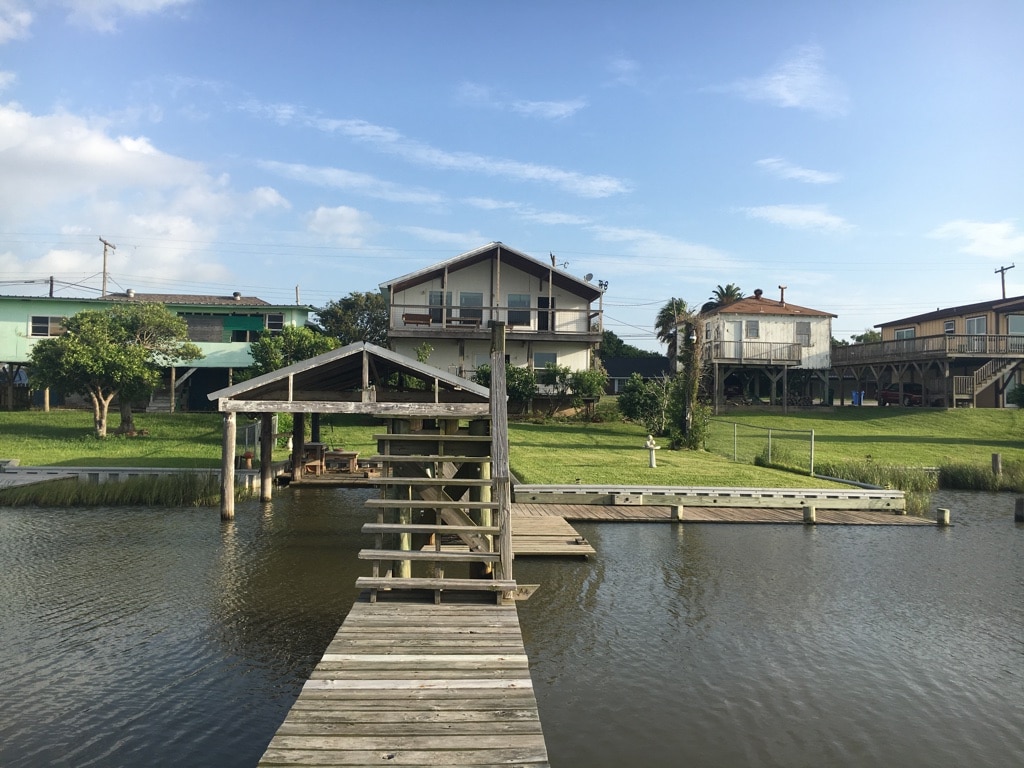 A wooden dock extends over calm waters, leading to a grassy area with two houses in the background. The houses feature off-white and green exteriors, with one elevated on stilts and surrounded by trees. A clear sky is visible, indicating a pleasant day.