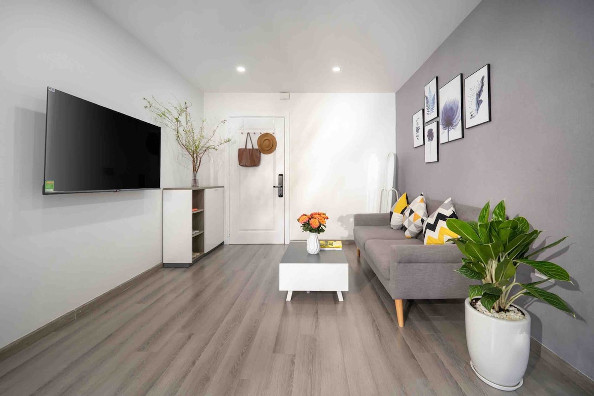 A modern living room features a spacious L-shape sofa adorned with decorative cushions. A coffee table sits at the center, accompanied by a blooming plant in a white pot. A wall-mounted TV is positioned opposite the sofa, with framed artwork displayed above.