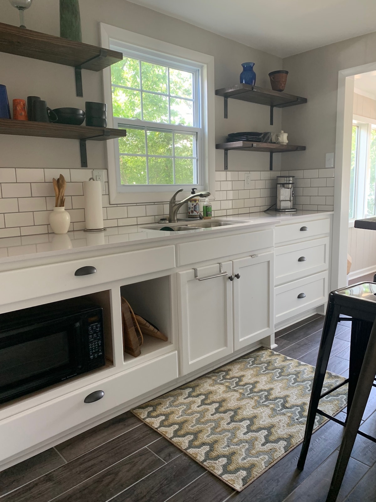 A modern kitchen is displayed, featuring white cabinetry, open shelving, and a stylish backsplash. A sink and coffee maker are visible, complemented by a microwave tucked in a cabinet. Natural light enters through a window, illuminating a patterned rug situated on the floor.