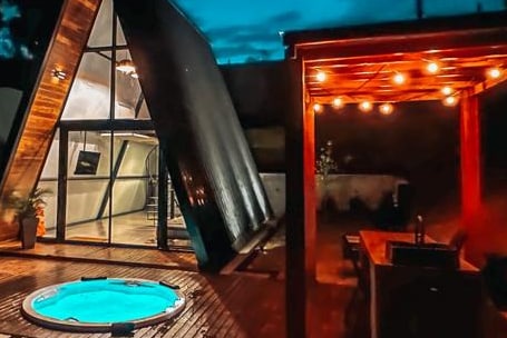 The image depicts a modern cabin with a distinct triangular shape, featuring large glass windows. An outdoor deck includes a private hot tub, a dining area with wooden furnishings, and string lights providing soft illumination, creating a calm ambiance for relaxation.