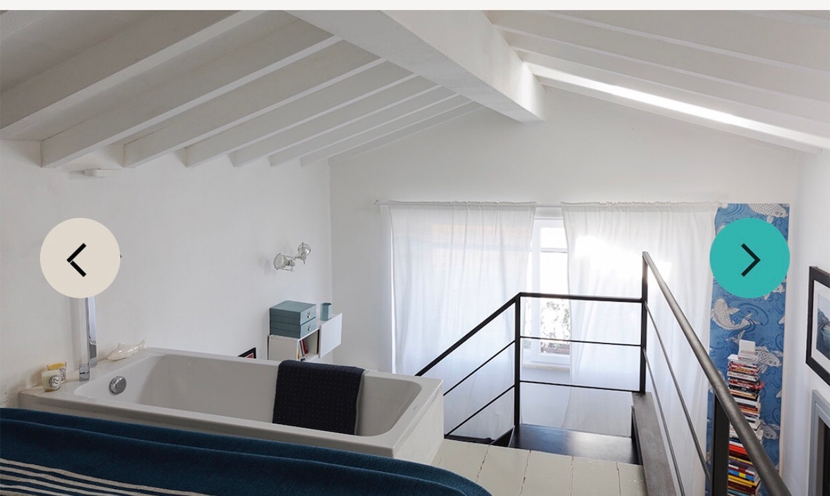 An elevated view of a spacious loft showcasing a freestanding tub, framed by white walls and wooden beams. A large window allows natural light to illuminate the area, complemented by sheer curtains. The design includes a sleek railing that overlooks the lower level.