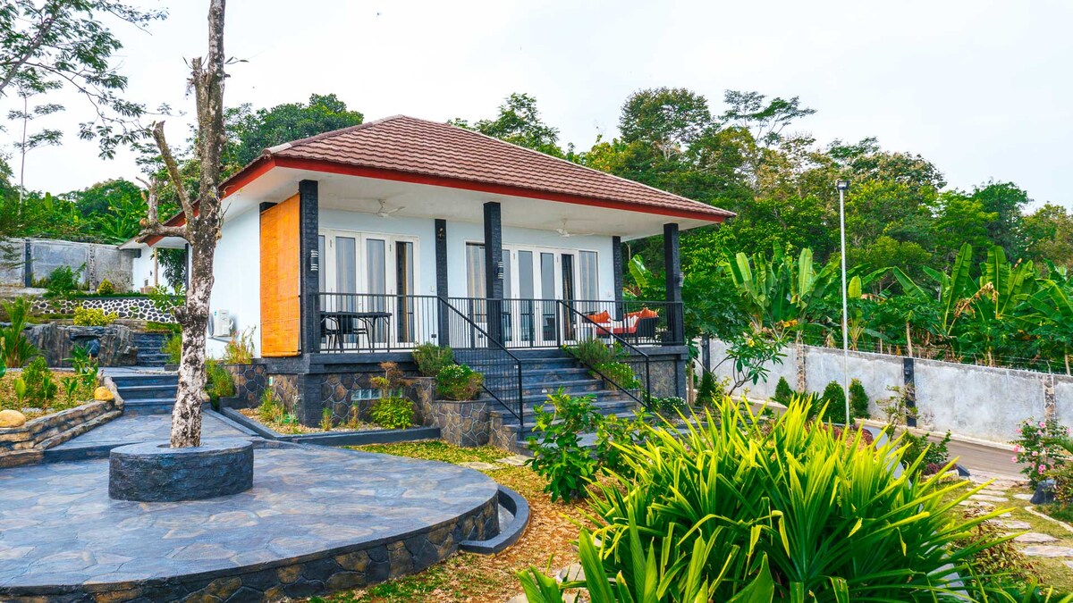 The standalone villa is framed by lush greenery, featuring a modern exterior with a sloping roof. Large glass doors provide access to a spacious patio area, which is furnished for outdoor relaxation. The surrounding landscape includes well-maintained gardens and stone pathways.