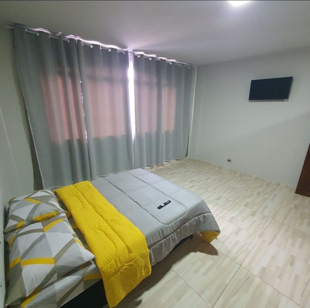 A spacious sleeping area features a neatly made bed with a gray and yellow blanket. Soft natural light filters through light gray curtains, while a small wall-mounted TV is visible on the opposite wall.