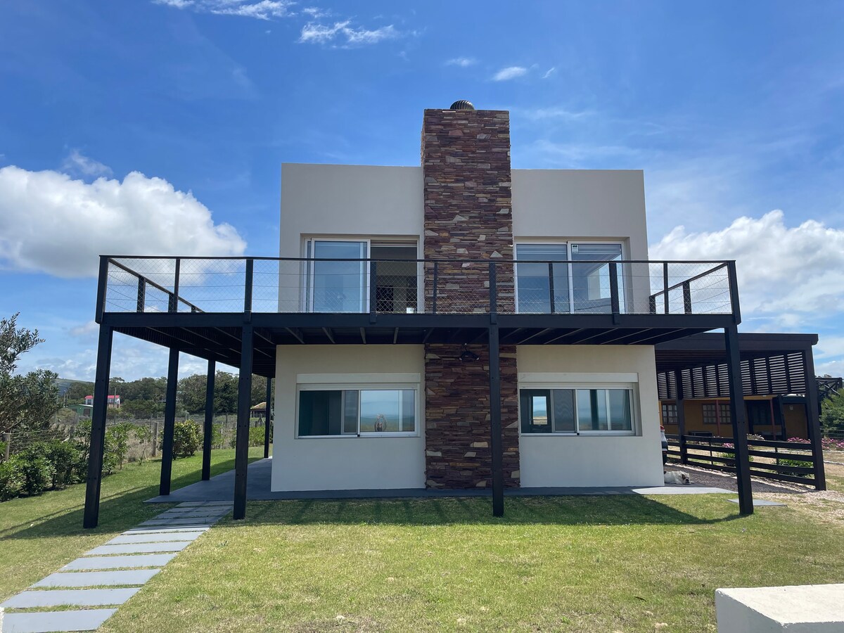 The two-story house features a modern design with a prominent stone chimney. A large deck extends over the entrance, providing panoramic views of the surrounding natural landscape. Lush greenery and a pathway lead to the front, while large windows enhance the home's contemporary aesthetic.