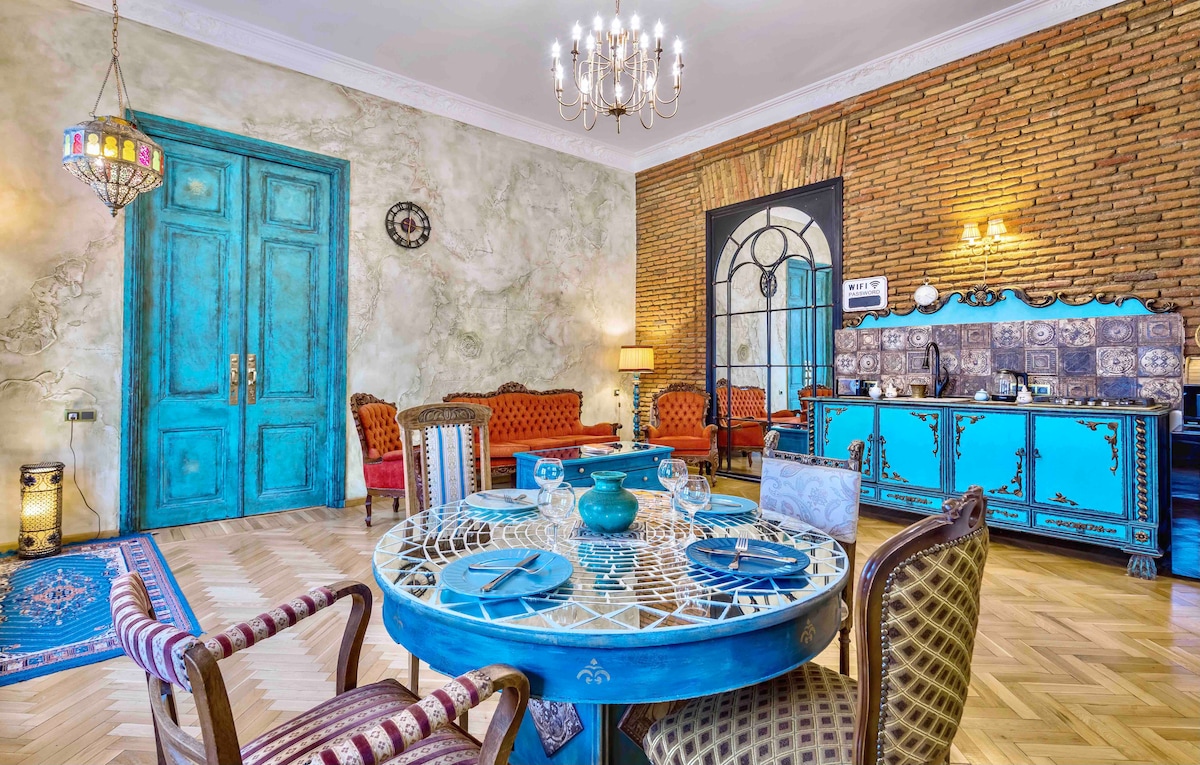 A vibrant dining area features a decorated table set with dishes and a centerpiece. Antique chairs add charm to the space, accented by a large blue cabinet and warm lighting. The textured walls and decorative elements create a visually engaging atmosphere.