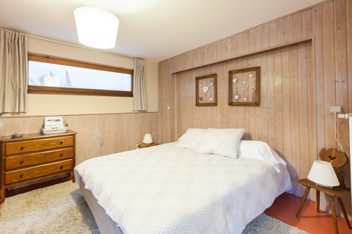 A sleeping area features a spacious bed with white bedding, complemented by a light wooden headboard. Natural light enters the room through a window dressed with light curtains. A bedside table and a wooden dresser add functional touches to the neat space.
