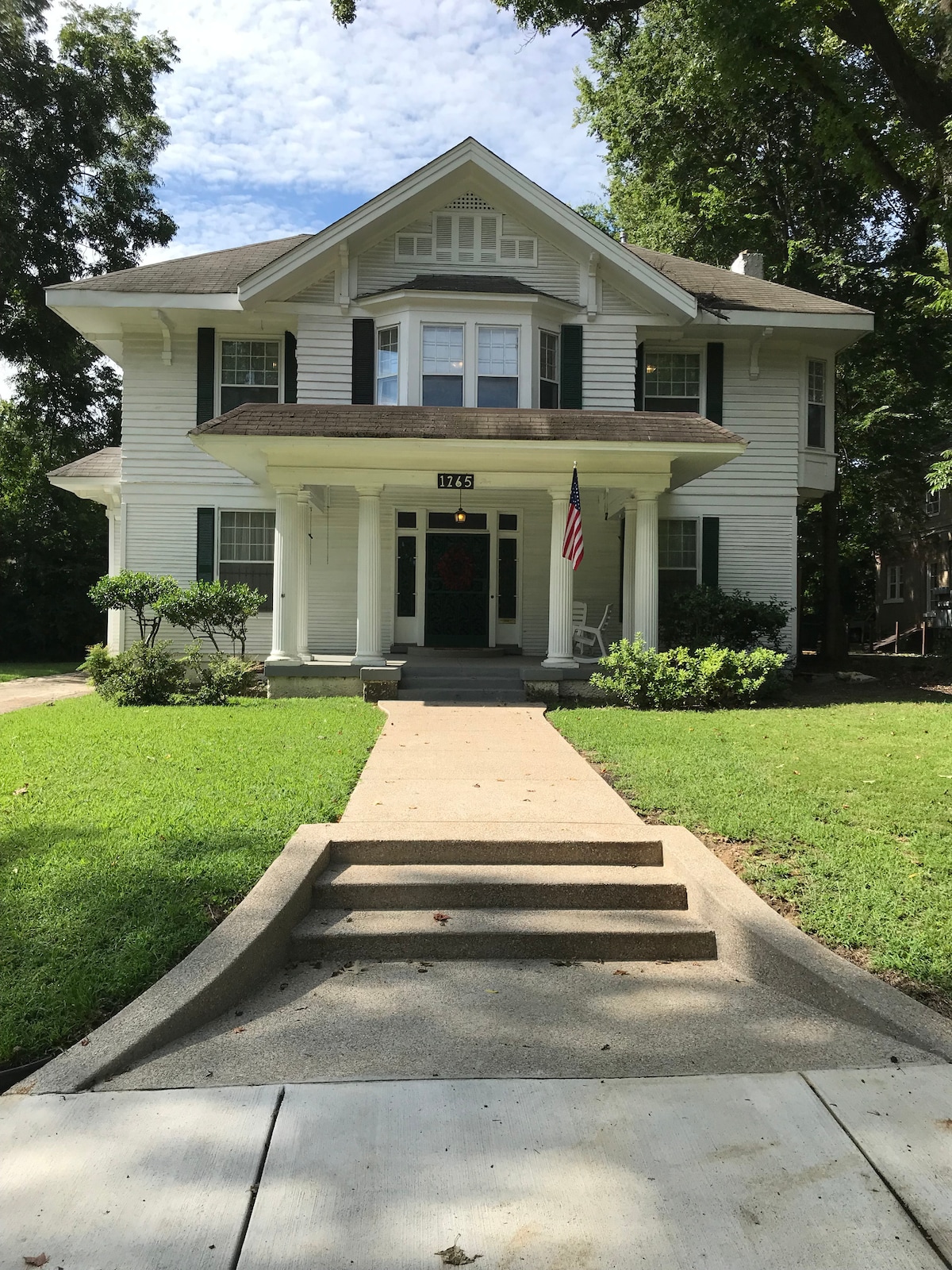 A historic two-story home with white siding and green shutters is surrounded by well-maintained landscaping. Steps lead up to a covered porch, adorned with a wreath. An American flag hangs from a front column, contributing to the home's classic charm.