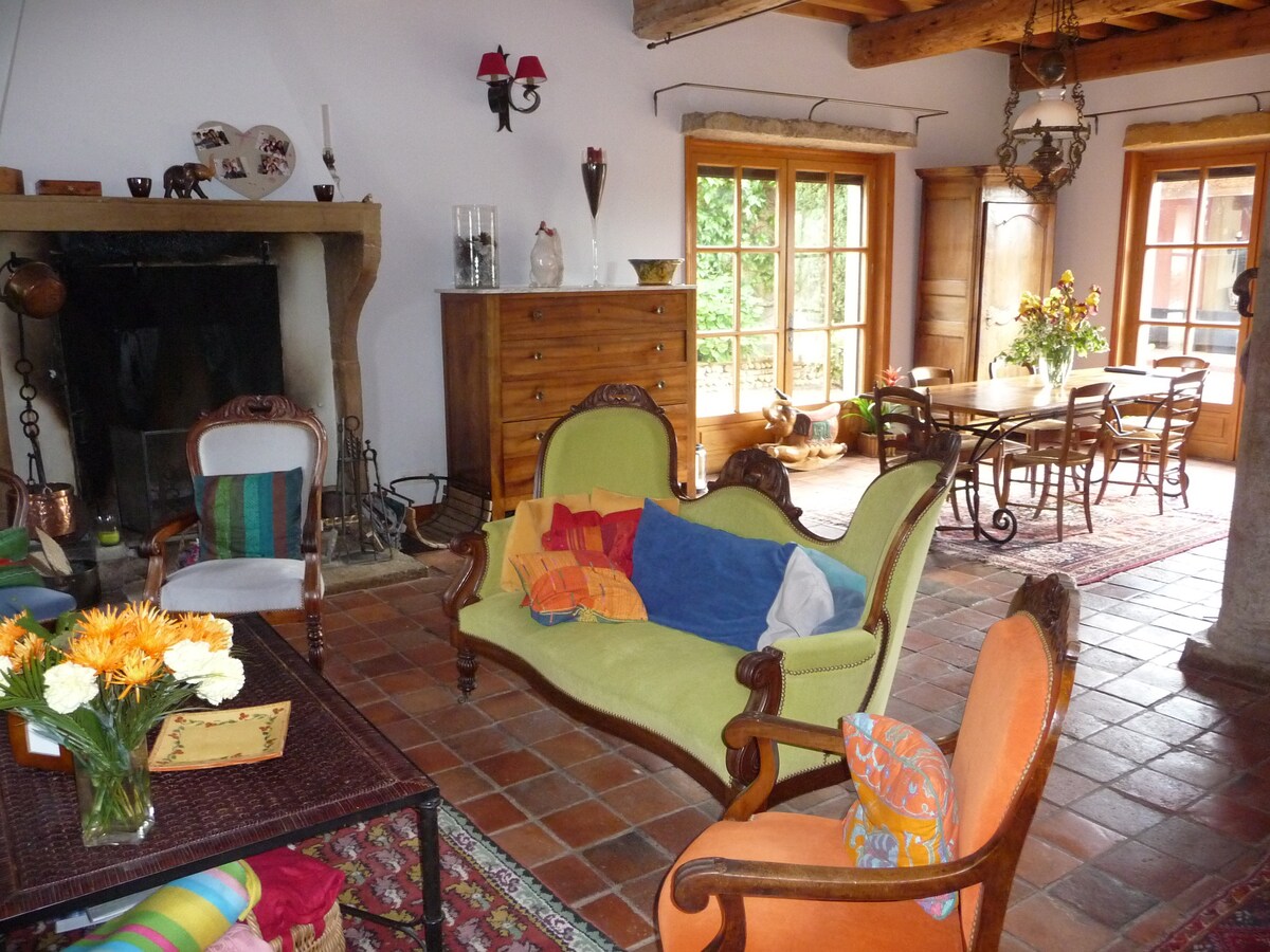 A charming living area features a large, green upholstered sofa accompanied by colorful cushions. An antique wooden cabinet stands against the wall, and a dining table with chairs is visible in the background. Natural light floods the space through multiple framed windows.