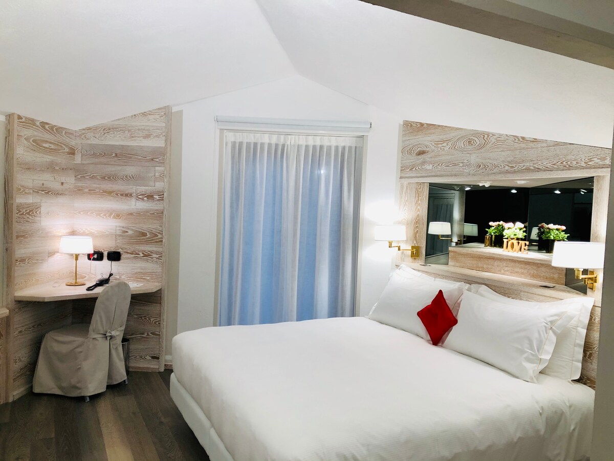 A well-arranged sleeping area features a spacious bed with white linens and a decorative red pillow. Natural light filters through sheer curtains, and a desk is positioned nearby. Wooden accents create a warm ambiance, complemented by bedside lamps and a framed display of flowers.