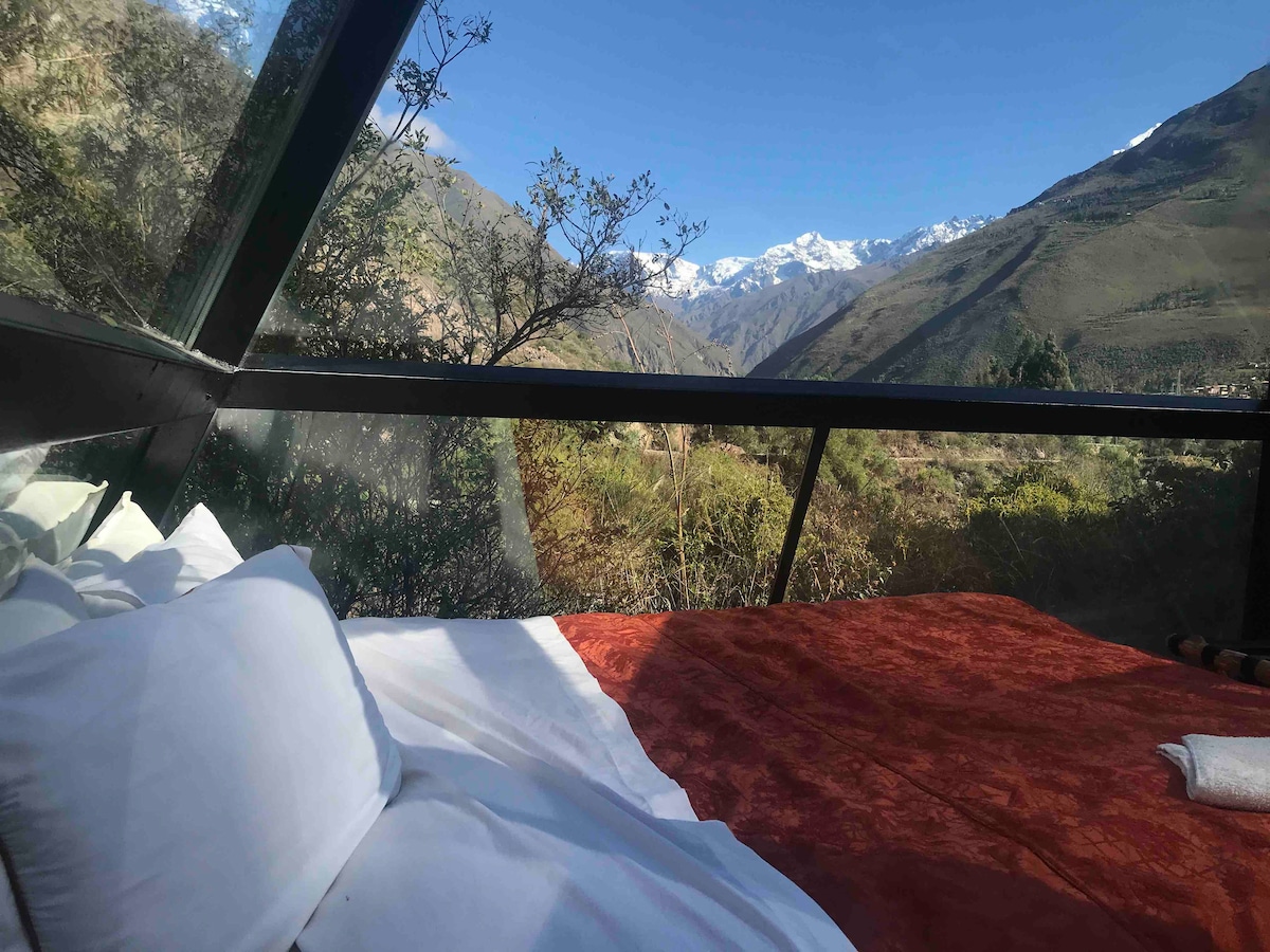 A comfortable bed, dressed with soft linens, is positioned alongside expansive glass walls, offering views of the surrounding mountains and greenery. Natural light floods the space, creating a serene atmosphere that connects the indoors with the stunning outdoor landscape.