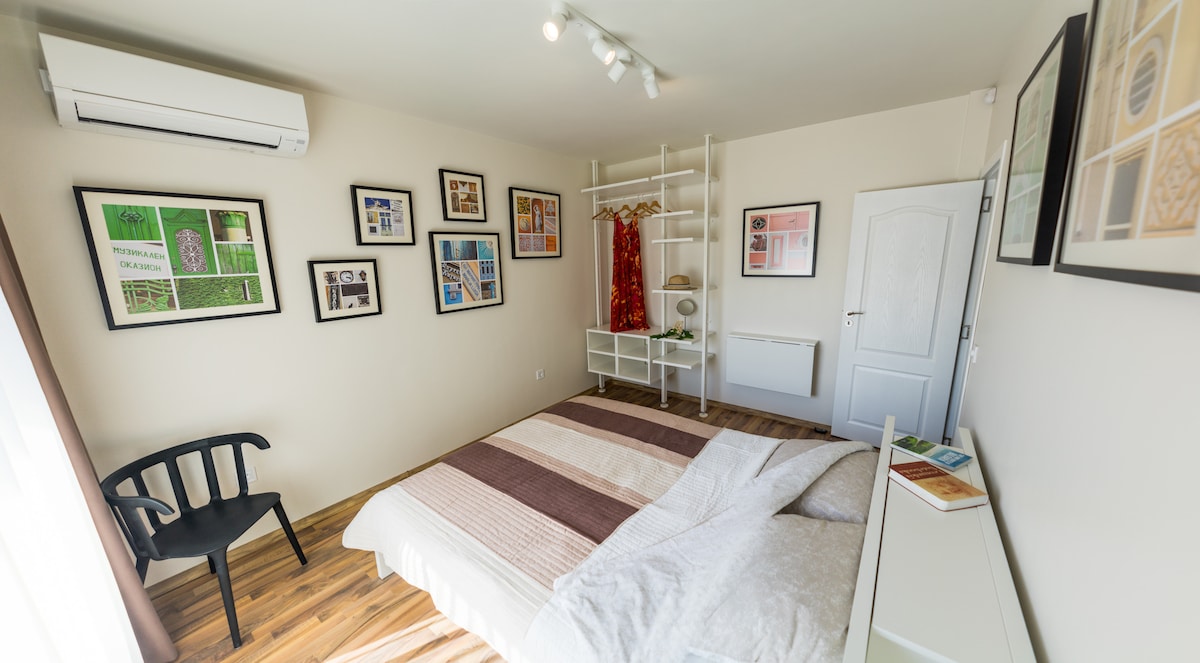 A comfortable bedroom features a spacious bed covered with light bedding, complemented by decorative wall art. A black chair is positioned beside a modern white shelf, and natural light streams in through large windows, enhancing the neutral color palette of the room.