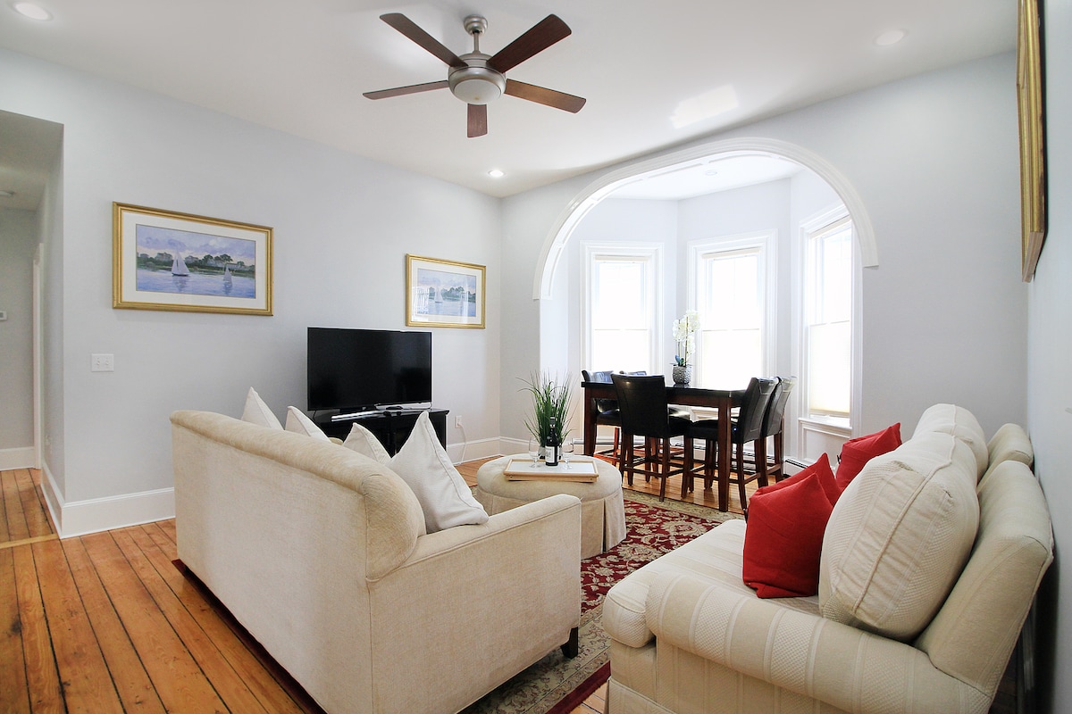 A bright living area features two sofas, one cream-colored with cushions and a red accent pillow. A television is positioned on a stand opposite the seating. Large windows provide natural light, complementing a dining area with dark wooden chairs and a table in the background.