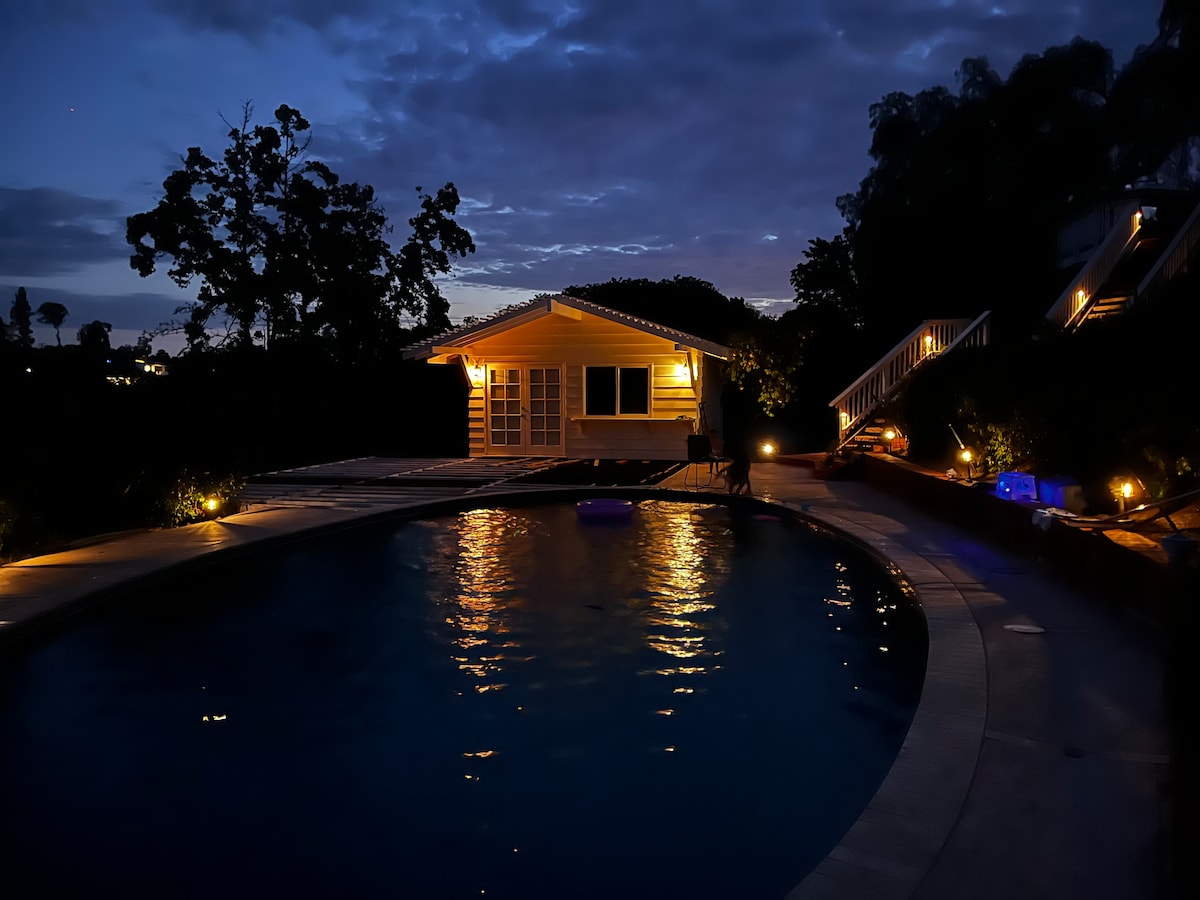 A serene view of a private pool surrounded by softly lit pathways, with a guest unit visible in the background. The water reflects the evening sky, while the landscape is gently illuminated by warm lights, enhancing the tranquil setting.