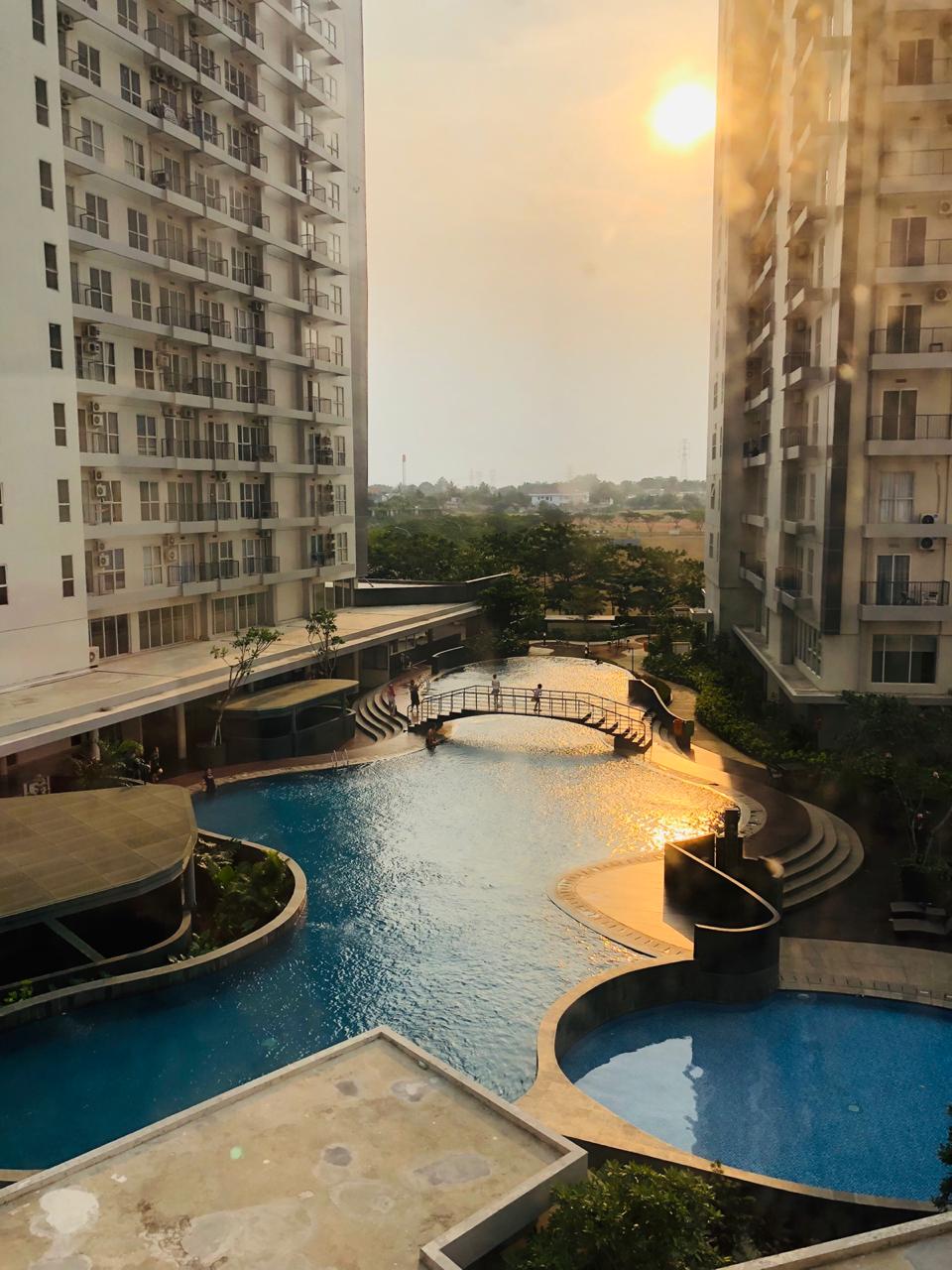 A scenic view of the swimming pool area is presented, with shimmering water reflecting the sunlight. Lounge spaces and a bridge are visible around the pool, framed by the modern architectural features of the surrounding buildings.