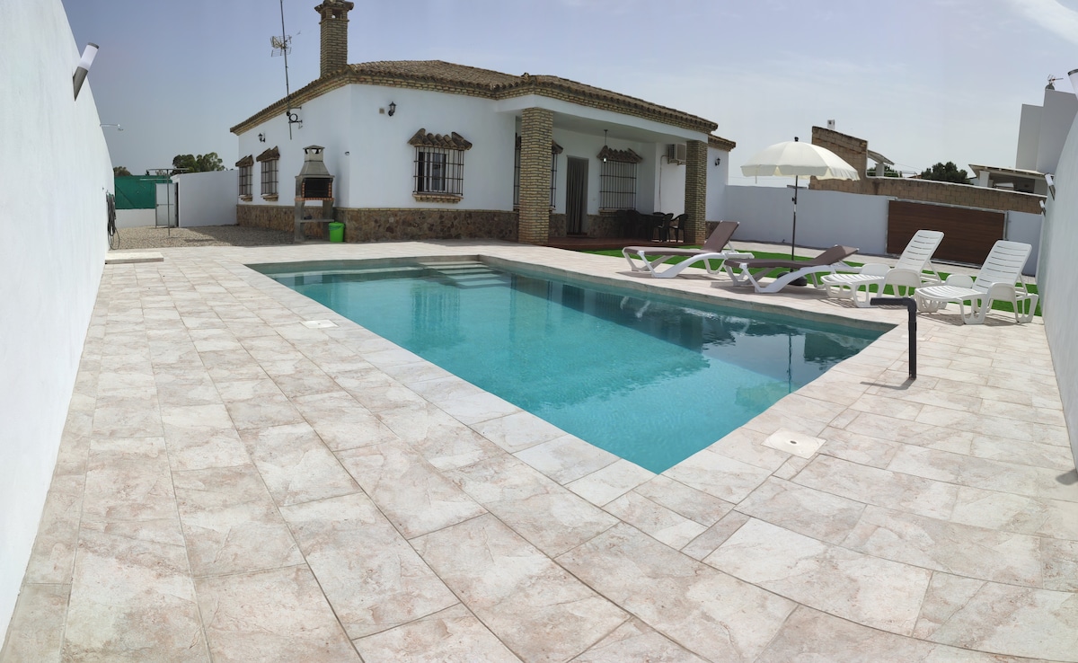 A spacious outdoor area features a large swimming pool surrounded by tiled decking. Lounge chairs are positioned alongside the pool, and an umbrella provides shade. A well-maintained stone house is visible in the background, contributing to a relaxing atmosphere.
