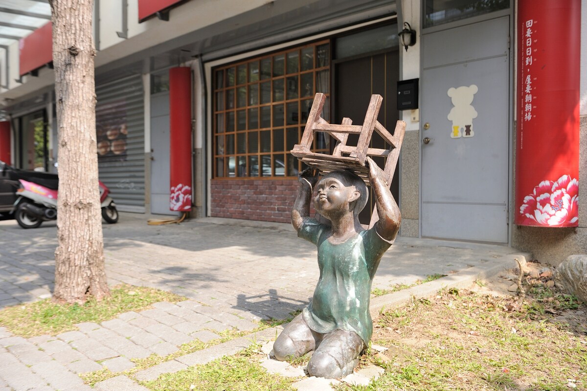 A bronze sculpture depicts a figure kneeling with a chair held above their head, situated on a landscaped area with pavers and grass. The background includes a storefront with large windows and red accents, contributing to the urban setting.