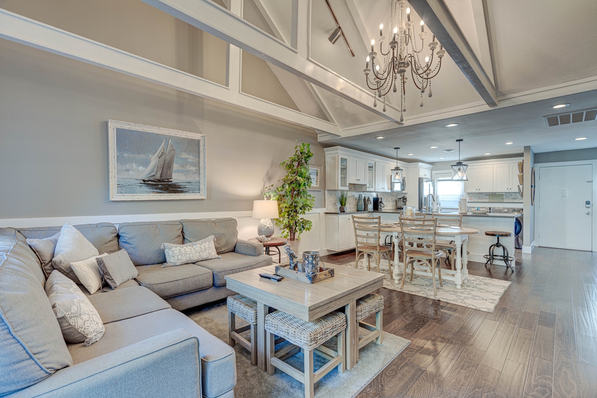 An inviting living space features a sectional sofa, a wooden coffee table, and a dining area with several chairs. Natural light illuminates the room through multiple windows, highlighting the coastal-inspired decor and airy layout. A chandelier complements the open, relaxed atmosphere.