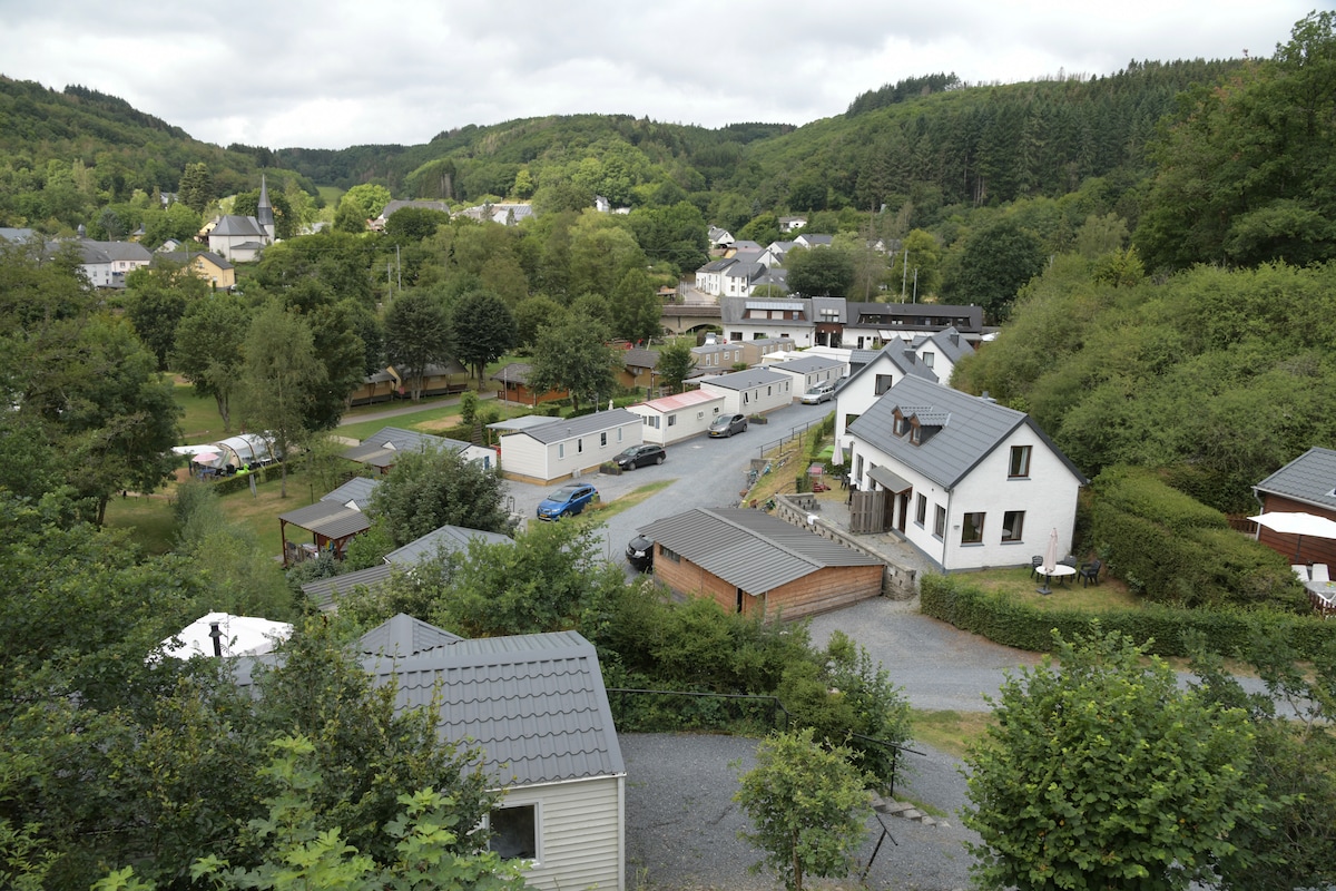 A view of the surrounding area captures a mix of residential structures and green spaces, nestled among hills. The image depicts several buildings, including a two-story home and nearby mobile homes, set along a winding road with trees and vegetation enhancing the landscape.