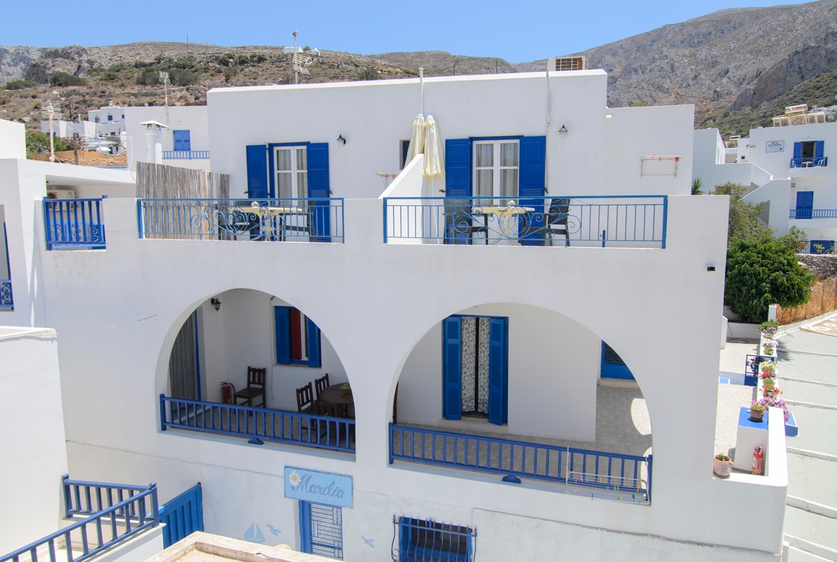 The exterior of the building is characterized by vibrant blue accents surrounding white walls. Arched balconies are visible, each furnished with chairs and a table. A sun umbrella offers shade in the outdoor space, enhancing the inviting atmosphere of the property.