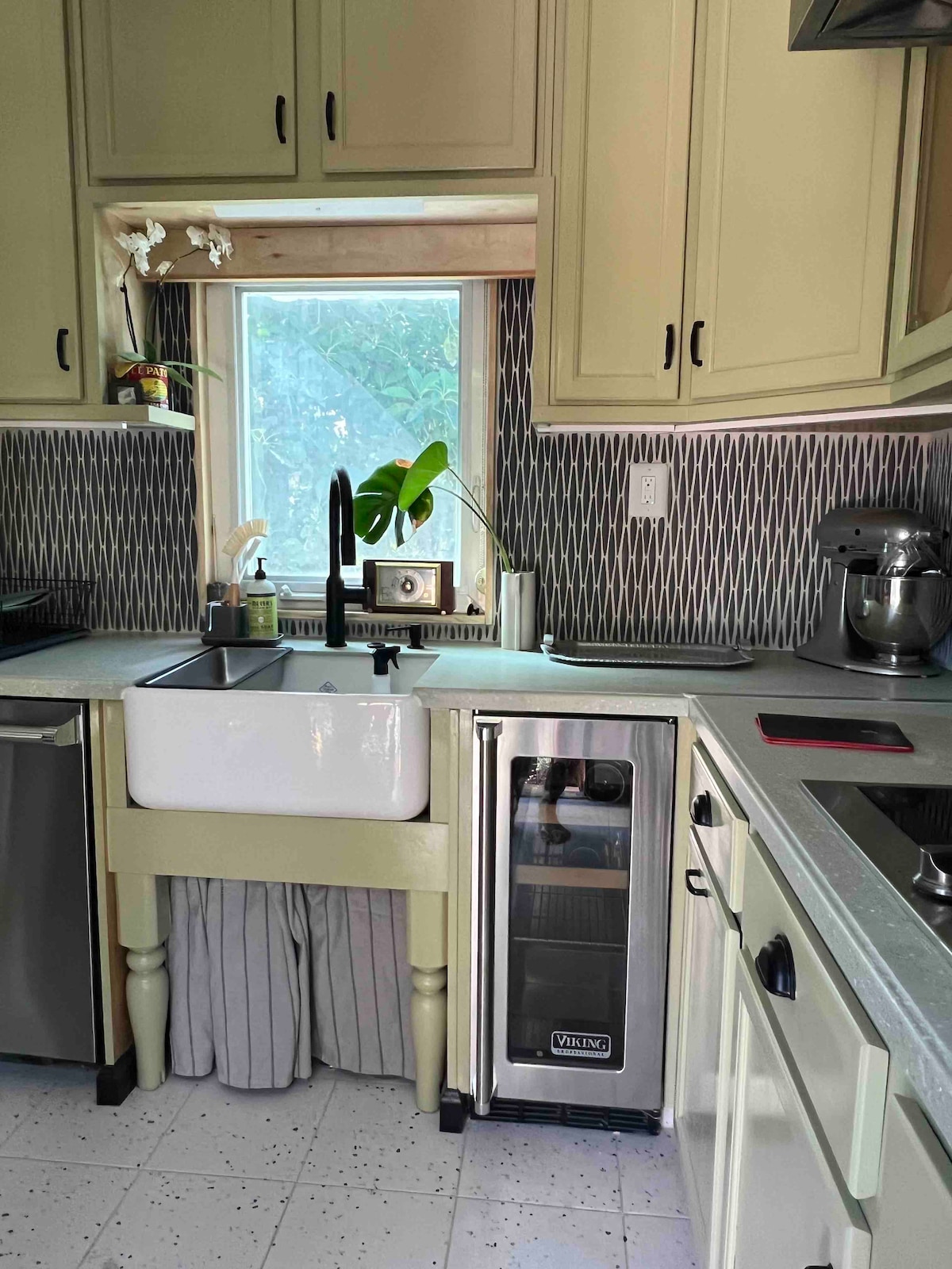 A functional kitchen space is showcased with cream-colored cabinetry and a farmhouse sink beneath a window. A VIKING wine fridge is positioned beside the sink, complementing the modern appliances. The counter features a variety of utensils and a plant, adding a touch of greenery.