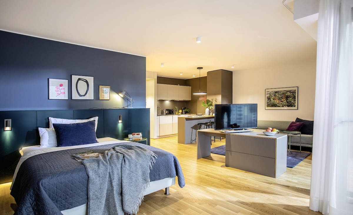 A spacious studio apartment is shown, featuring a comfortable bed with a textured blanket. Modern furnishings include a sleek dining table and a cozy seating area. A well-equipped kitchen is visible in the background, with stylish cabinetry and appliances. Natural light fills the room through large windows.
