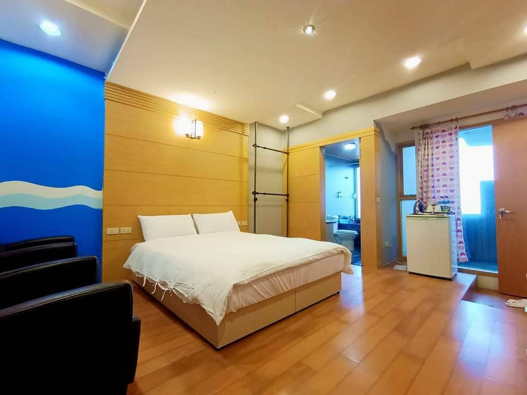 A spacious room features a large bed covered with a simple white blanket, flanked by two black lounge chairs. Light wood paneling complements blue accent walls. An ensuite bathroom is visible in the background, with a doorway leading to the kitchenette area adorned with patterned curtains.