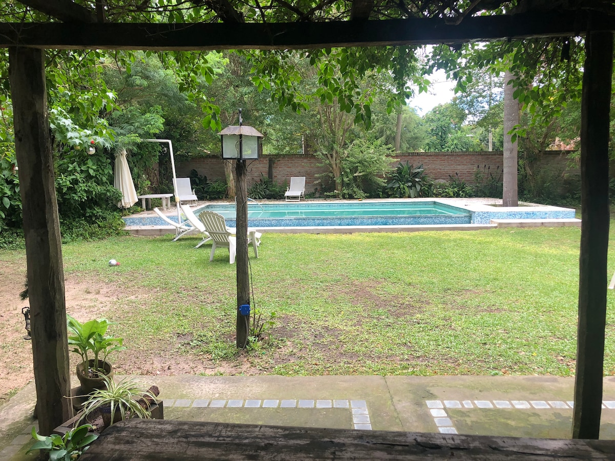 A view of a serene backyard reveals a swimming pool surrounded by lush greenery. Two lounge chairs are positioned beside the pool, and a shaded area provides a relaxing spot under leafy vines. The well-maintained lawn adds to the inviting outdoor space.