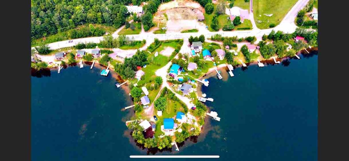 An aerial view captures a shared campground setting along Lake Nosbonsing, featuring multiple cabins surrounded by greenery. Docks extend into the water along the shoreline, with boats visible. The layout includes access paths and communal areas, indicating a vibrant outdoor space for guests.
