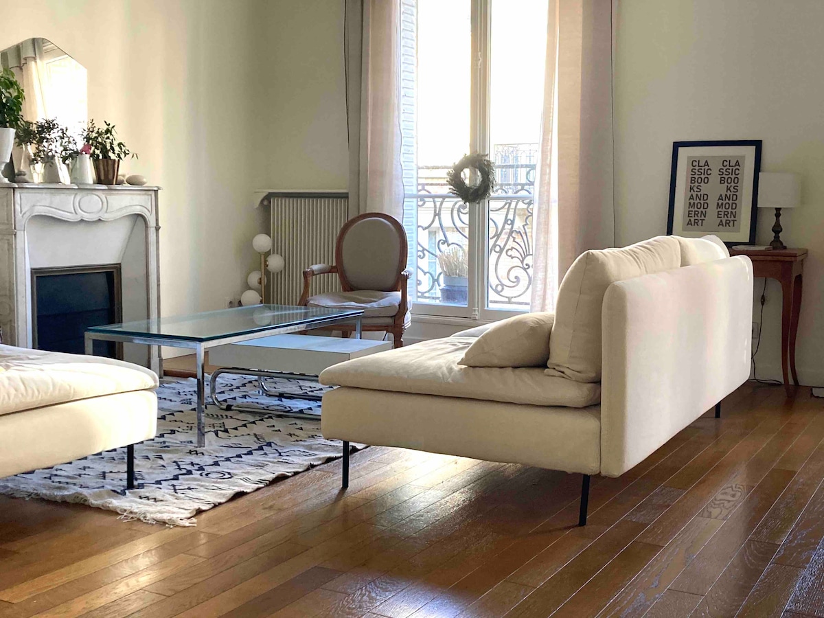 A bright living area features a light-colored sectional sofa and a glass coffee table. A wooden side table holds a lamp, while a decorative chair adds seating. Natural light floods in through large windows adorned with sheer curtains, revealing glimpses of the charming balcony.