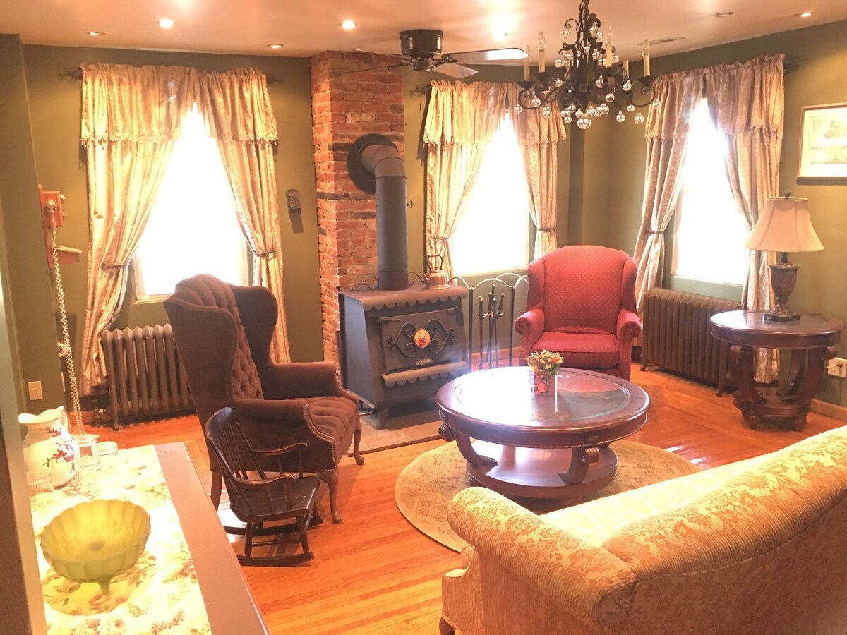 A cozy living area features large windows adorned with elegant drapes, allowing natural light to fill the space. A vintage wood-burning stove serves as a focal point, surrounded by comfortable seating options, including a red armchair and a rocking chair. A wooden coffee table sits in the center.