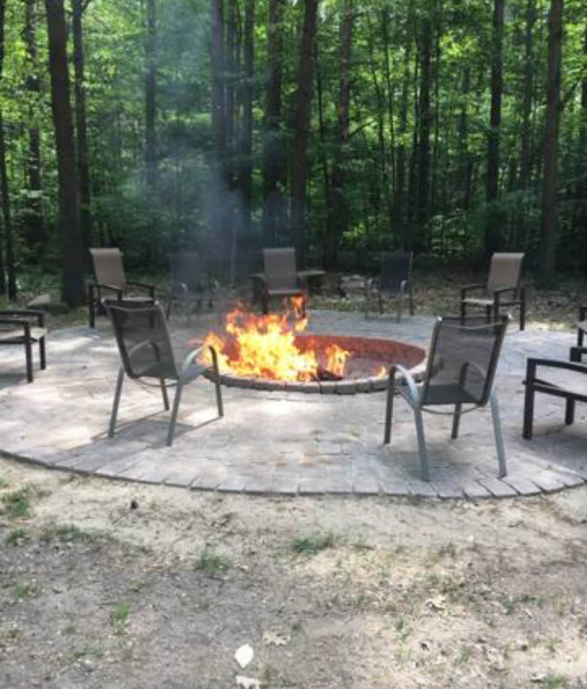A stone fire pit is surrounded by several chairs arranged in a circular formation. Flames are visible, providing warmth and light. Lush green trees form a natural backdrop, creating a serene outdoor gathering space.