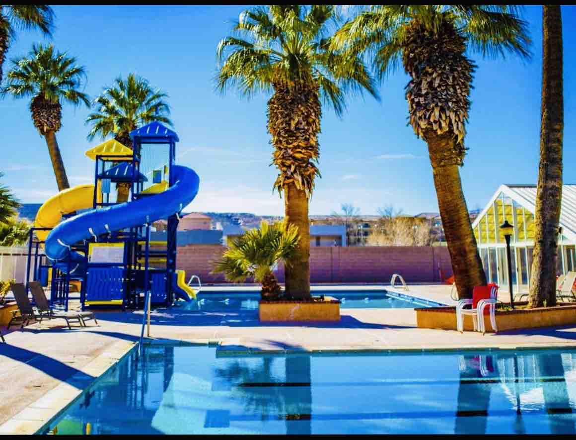A vibrant outdoor pool area features a yellow and blue water slide surrounded by tall palm trees. Lounge chairs are positioned around the pool, which reflects the clear blue sky. A separate shallow area for children is visible in the background.