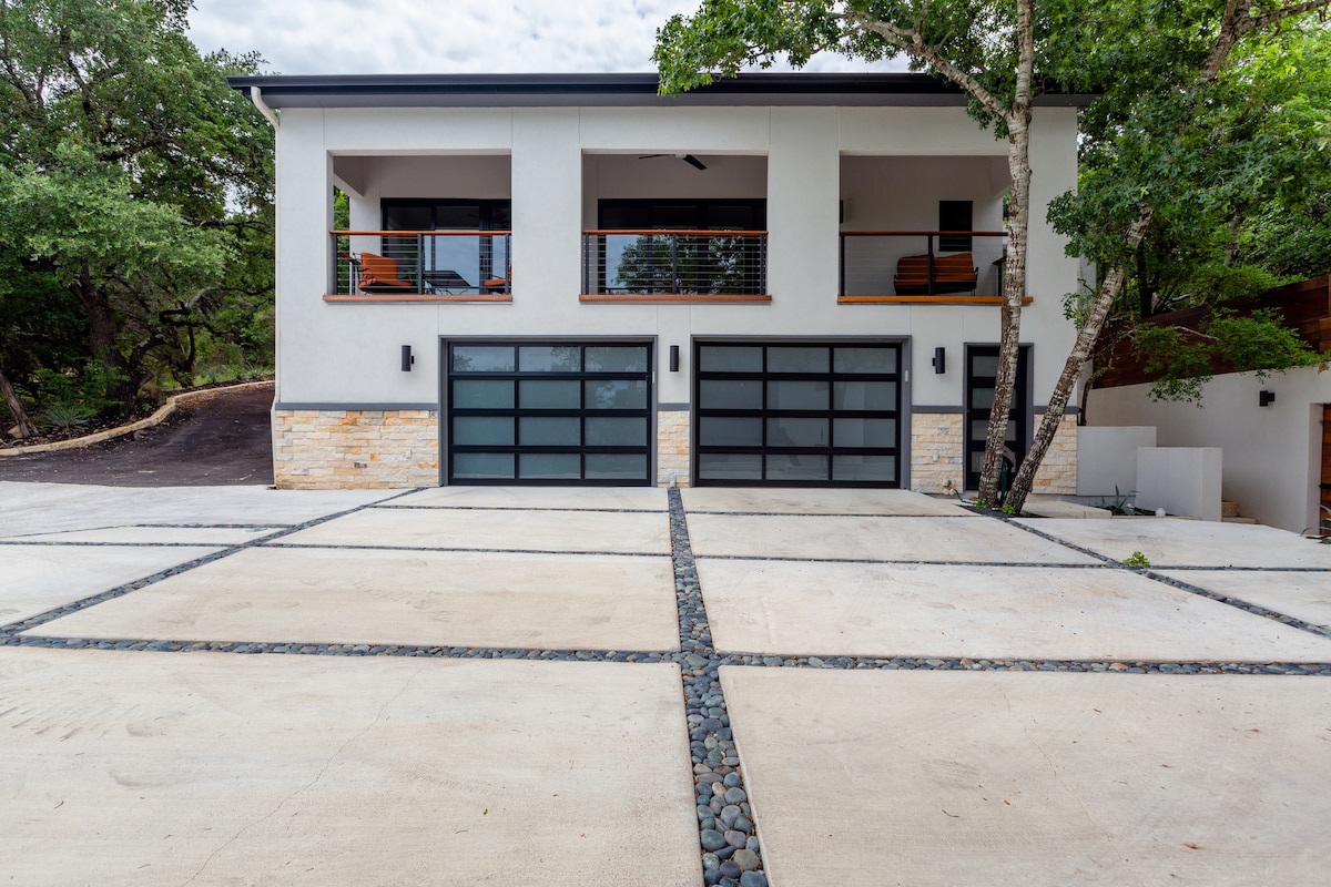 The exterior of the guest house is shown, featuring a modern two-story design with large glass garage doors. A spacious parking area is visible, with a stone pathway leading between the neatly arranged concrete slabs. Lush trees and greenery surround the property.