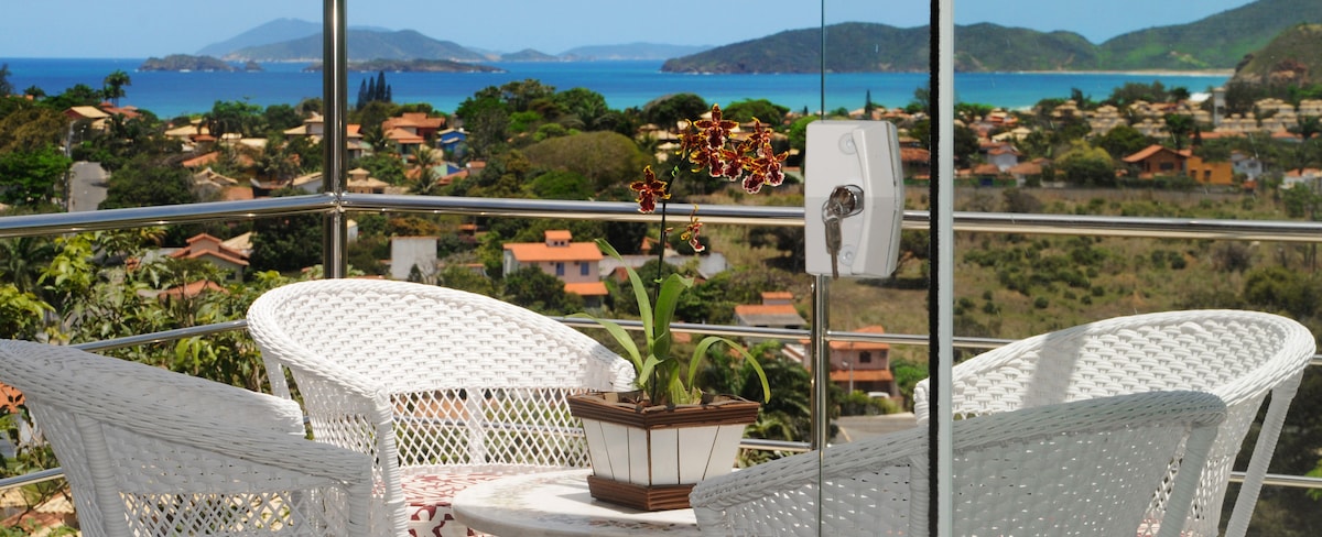 A balcony is shown with a round table and two white woven chairs. A small potted plant with flowers is placed at the center of the table. The background features a scenic view of the ocean and distant hills, with several houses visible in the foreground.