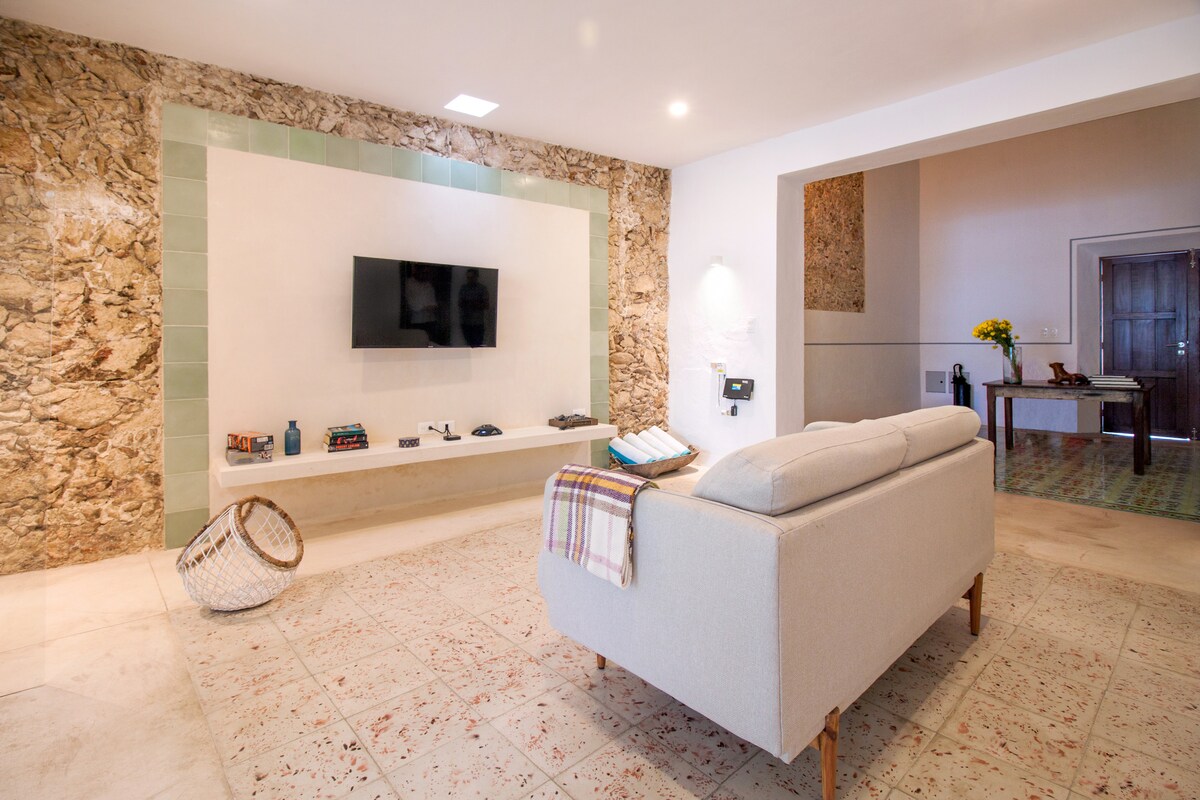 A modern living area features a light-colored sofa with a soft throw and a woven basket nearby. The room is anchored by a textured stone wall with an integrated TV. Natural light illuminates the space, creating a welcoming atmosphere for relaxation.