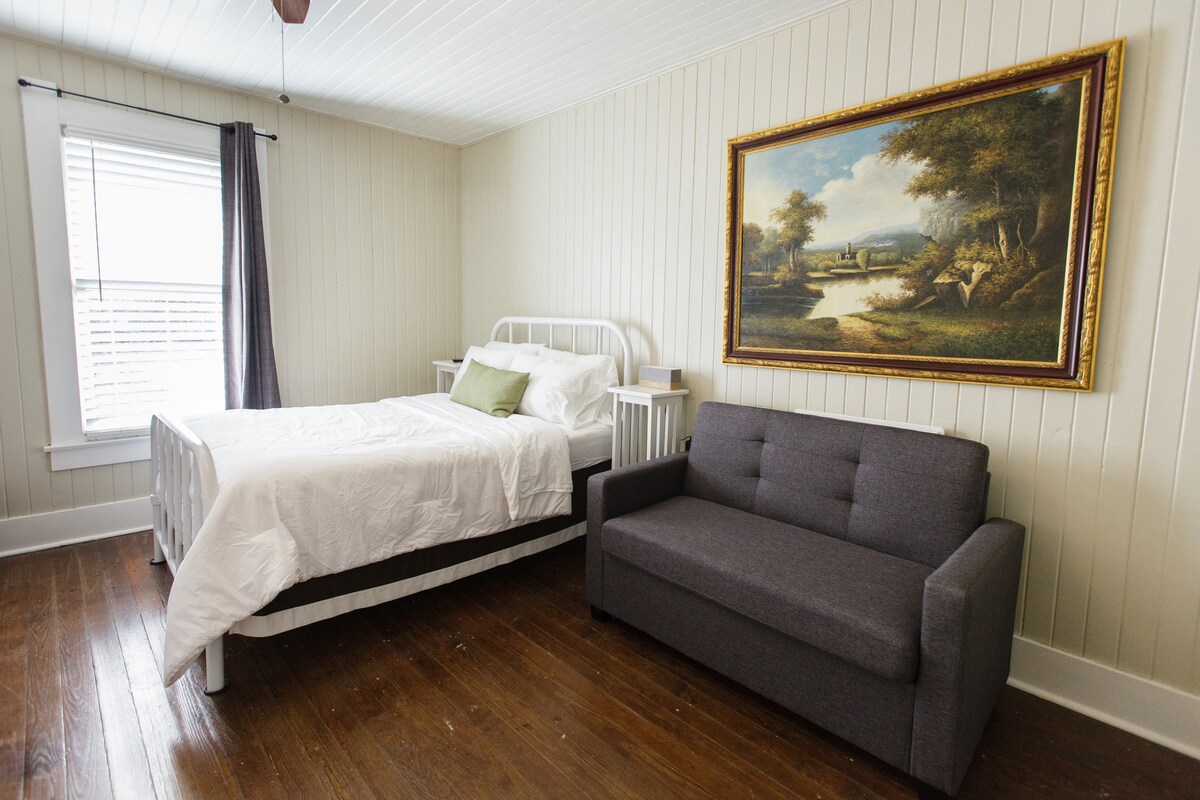 A bright sleeping area features a full bed with white bedding and a green accent pillow. A gray loveseat is positioned nearby, and a landscape painting adorns the wall. Natural light filters through a window covered by light curtains.