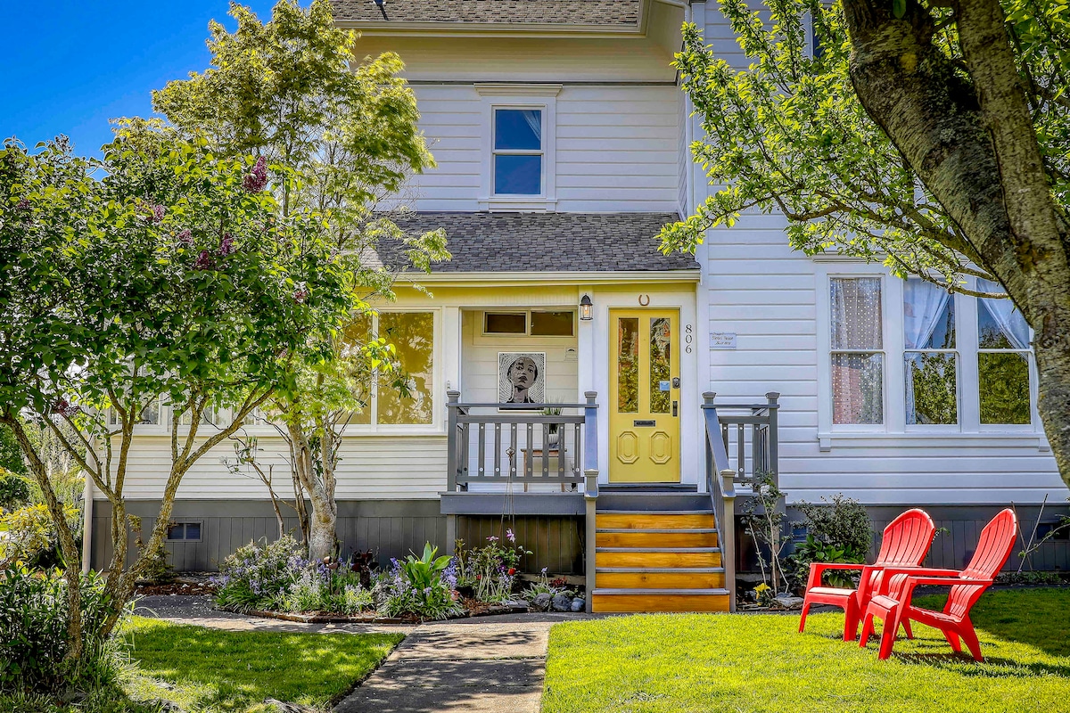 The charming entrance of a Victorian farmhouse features a bright yellow door and a welcoming porch. Red Adirondack chairs sit on the lawn, surrounded by manicured flower beds and greenery. The facade of the house showcases light-colored siding and large windows, allowing natural light to fill the interior.