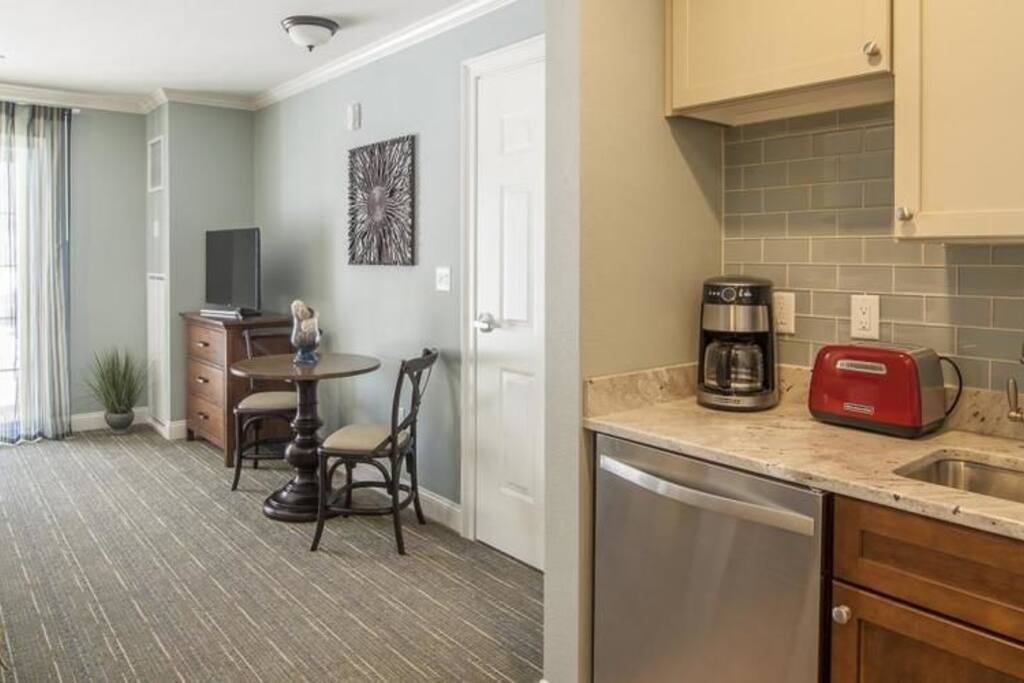View of TV, table and kitchen which has a small refrigerator, microwave, sink, & coffee maker.