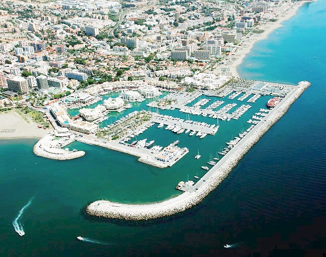 An aerial view of Puerto Marina in Benalmádena reveals a vibrant coastal area with numerous yachts docked in a well-organized marina. The surrounding beach and cityscape showcase a blend of lush greenery and modern architecture against the deep blue waters of the Mediterranean Sea.